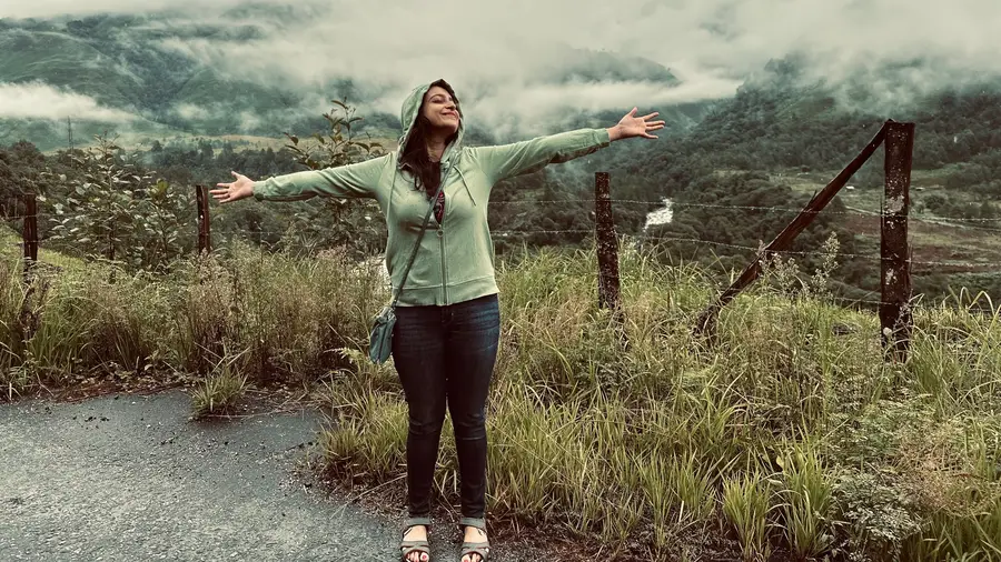 Megholova on a hillside road with arms outstretched, clouds rolling over the mountains behind