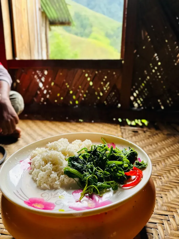 A plate of rice and cooked greens with king chilli on a woven bamboo floor, hills visible through the window