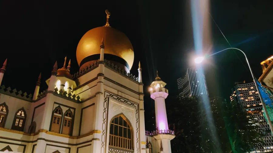 The golden dome and lit minaret of the Sultan Mosque at night, with office towers behind